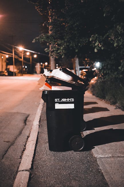 A black wheeled rubbish bin positioned on the edge of a street at night, with a white label bearing the name 'St. John's' on the front. The bin is overflowing with mixed waste, including paper, cardbo