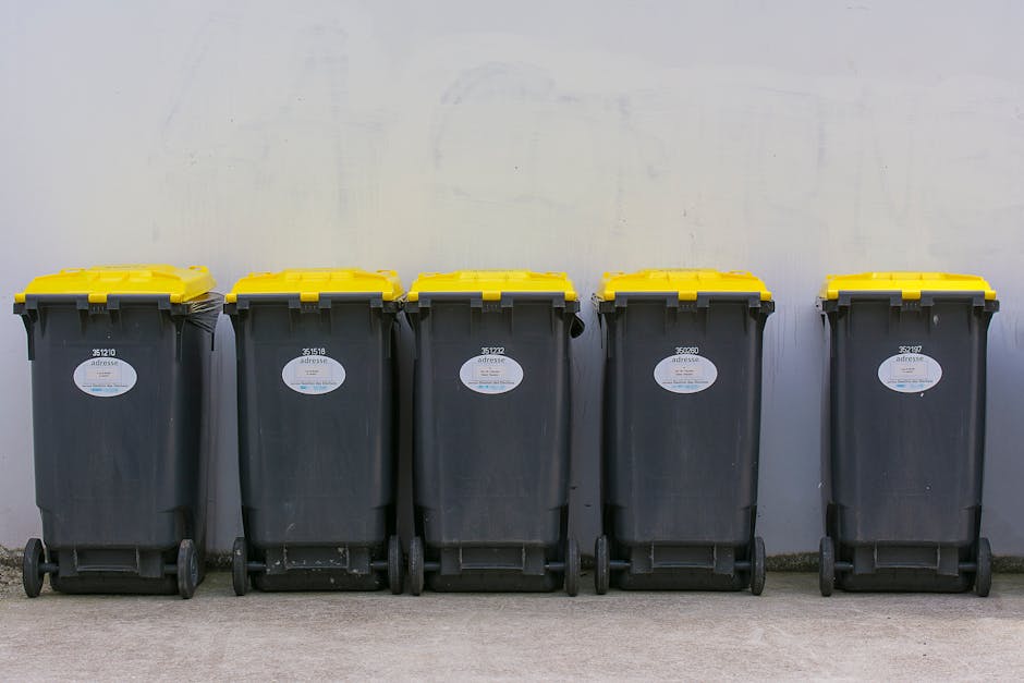 A black wheeled rubbish bin positioned on the edge of a street at night, with a white label bearing the name 'St. John's' on the front. The bin is overflowing with mixed waste, including paper, cardbo