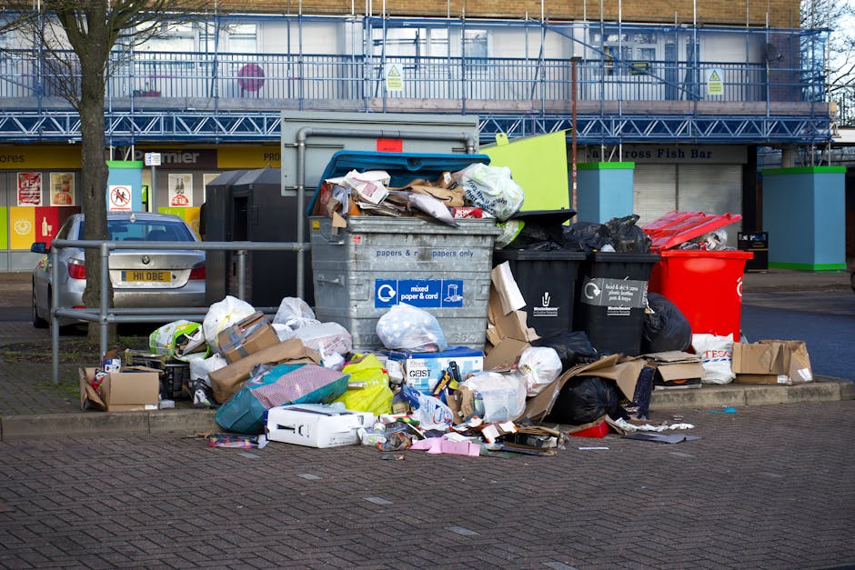 A narrow, outdoor alleyway lined with brick and metal walls, both covered in multicoloured graffiti. Various discarded items, including a white washing machine, a large cardboard box, a plastic bucket